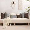 A sofa decorated with various textured gray and brown chenille throw pillow covers and a cream knit blanket.