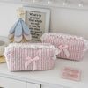 A pink and white striped cosmetic pouch with a lace trim is displayed on a white shelf next to a wooden rose decoration.