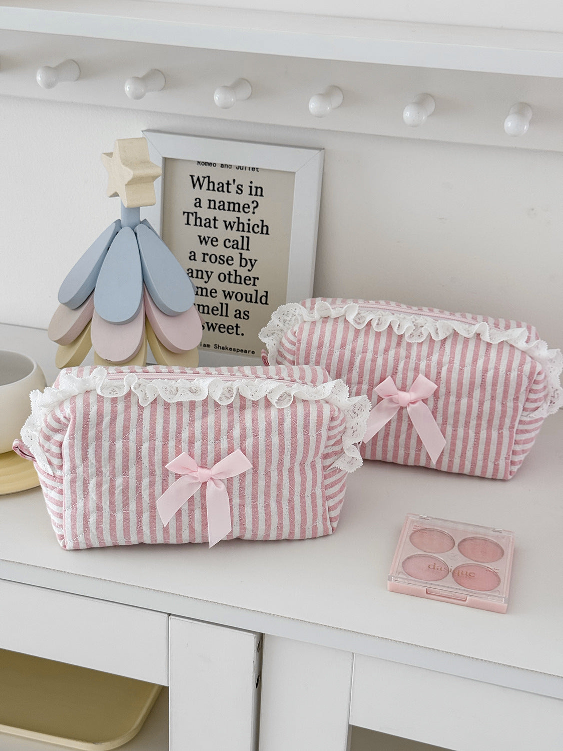 A pink and white striped cosmetic pouch with a lace trim is displayed on a white shelf next to a wooden rose decoration.