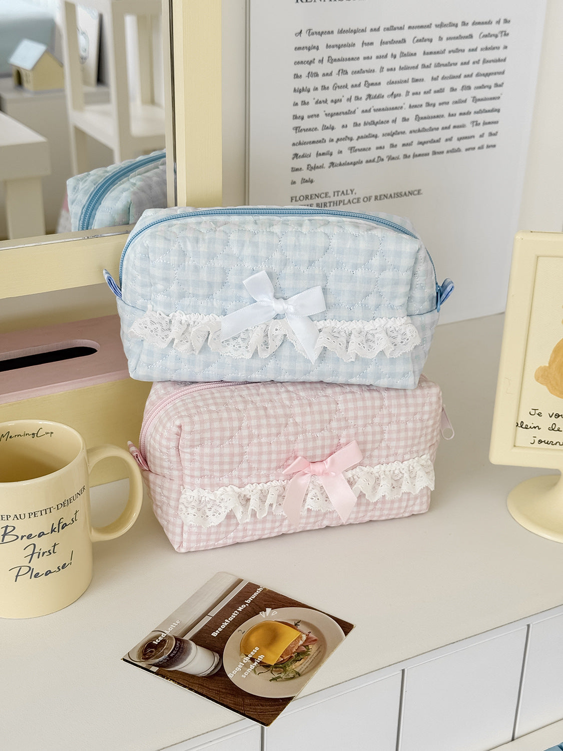 A light blue cosmetic pouch with white lace trim rests on a white surface next to a mug and a piece of food.