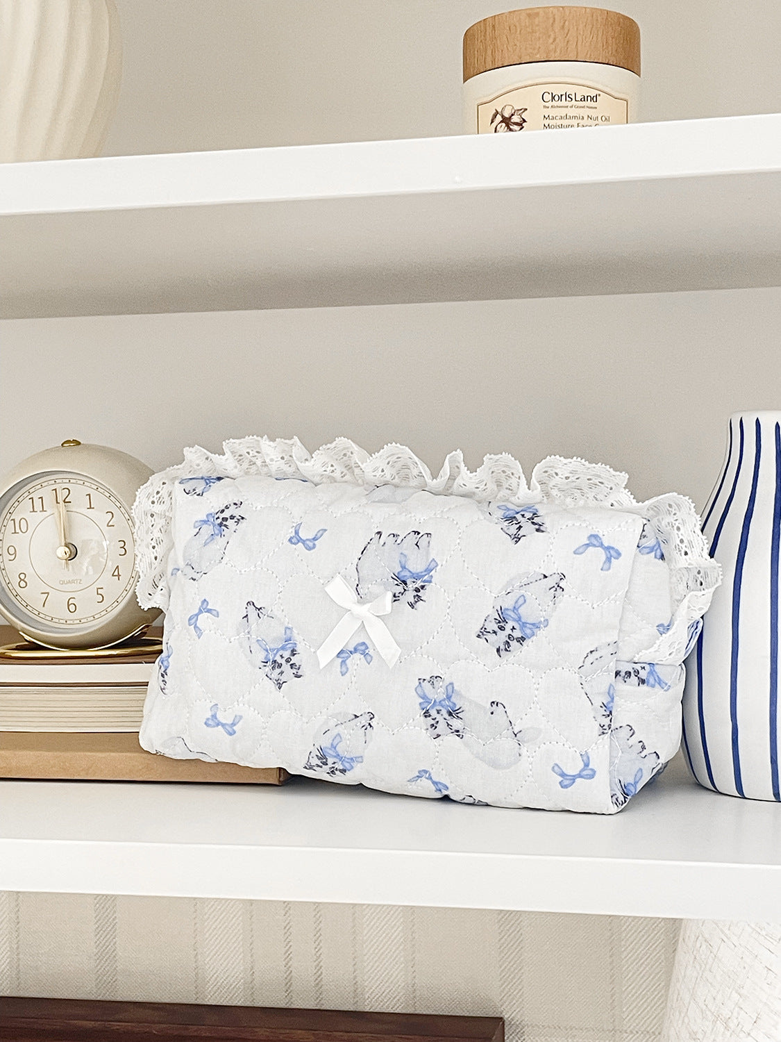 A white cosmetic pouch with a vintage rose print and lace trim resting on a white shelf.