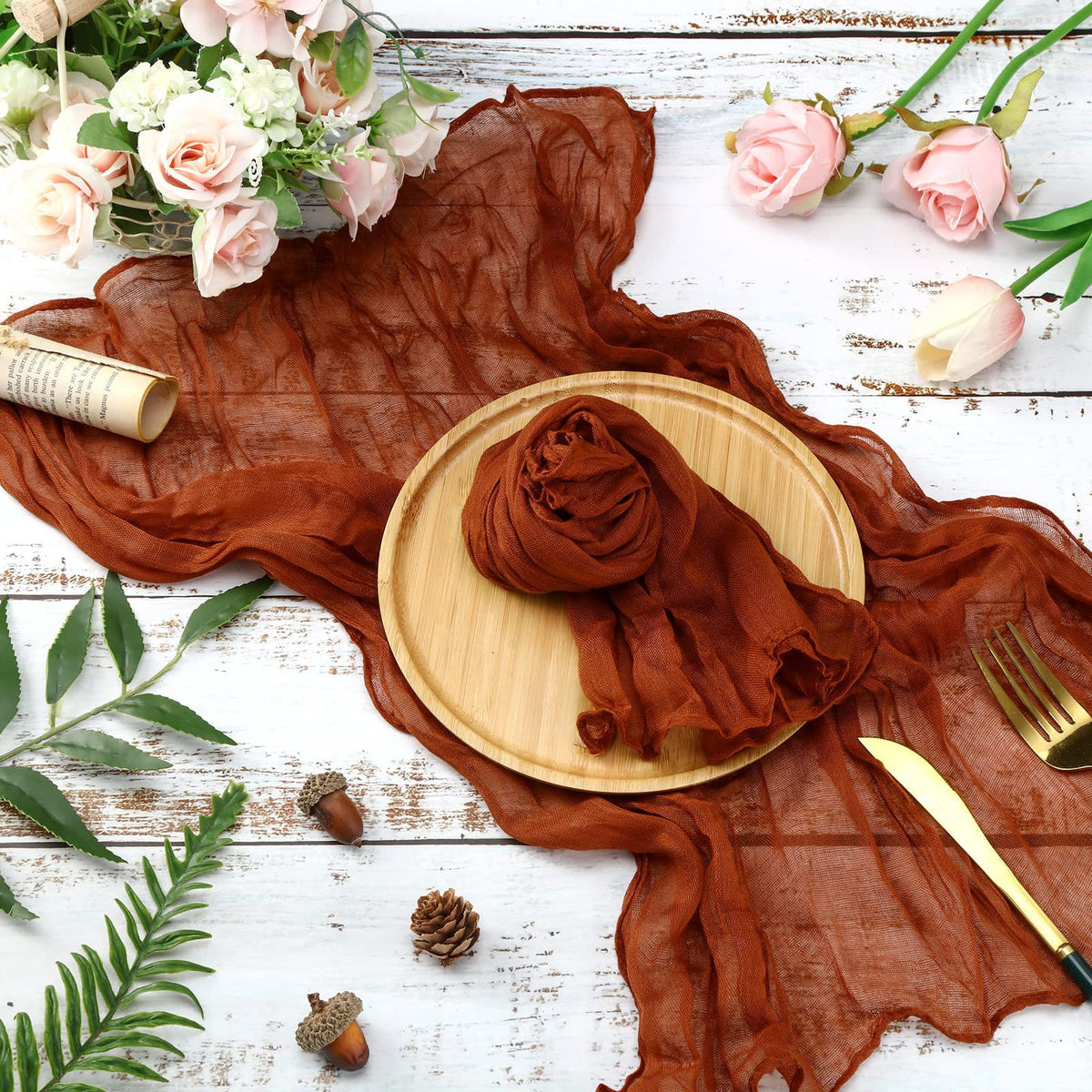 A rust brown cotton gauze table runner draped over a wooden plate on a white rustic surface.