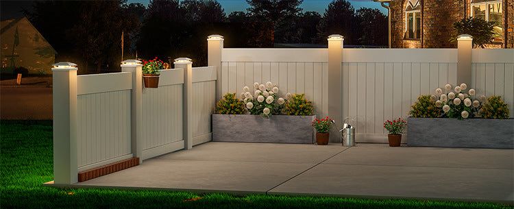 A white wooden fence illuminated by solar post cap lights with warm LED for a backyard patio.