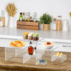 Stackable acrylic display risers used to elevate desserts and party buffet items on a wooden surface.