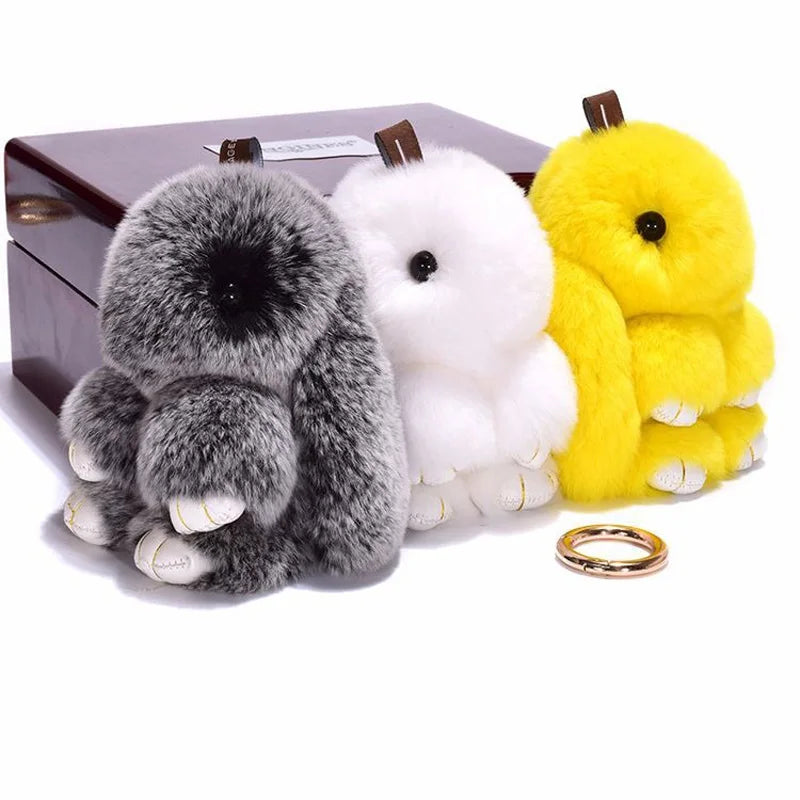 Three fluffy angel bunny keychains in gray, white, and yellow with pom pom tails displayed on a dark box.