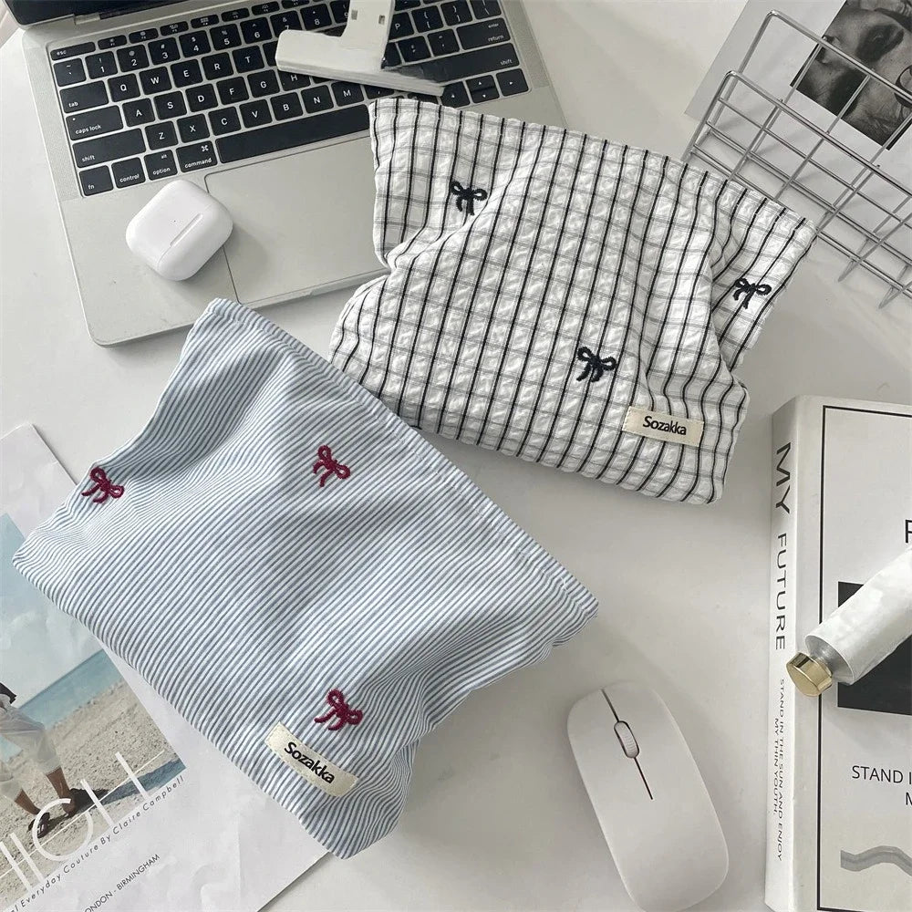 A light blue and white striped makeup bag with red floral embroidery is displayed next to a laptop and a white wireless mouse