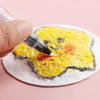 A hand applies black paint to a yellow puffy texture painting paper shaped like a smiling face.