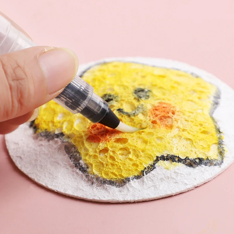 A hand applies black paint to a yellow puffy texture painting paper shaped like a smiling face.