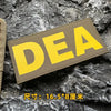 A yellow DEA embroidered patch is displayed on a dark, rough stone surface.