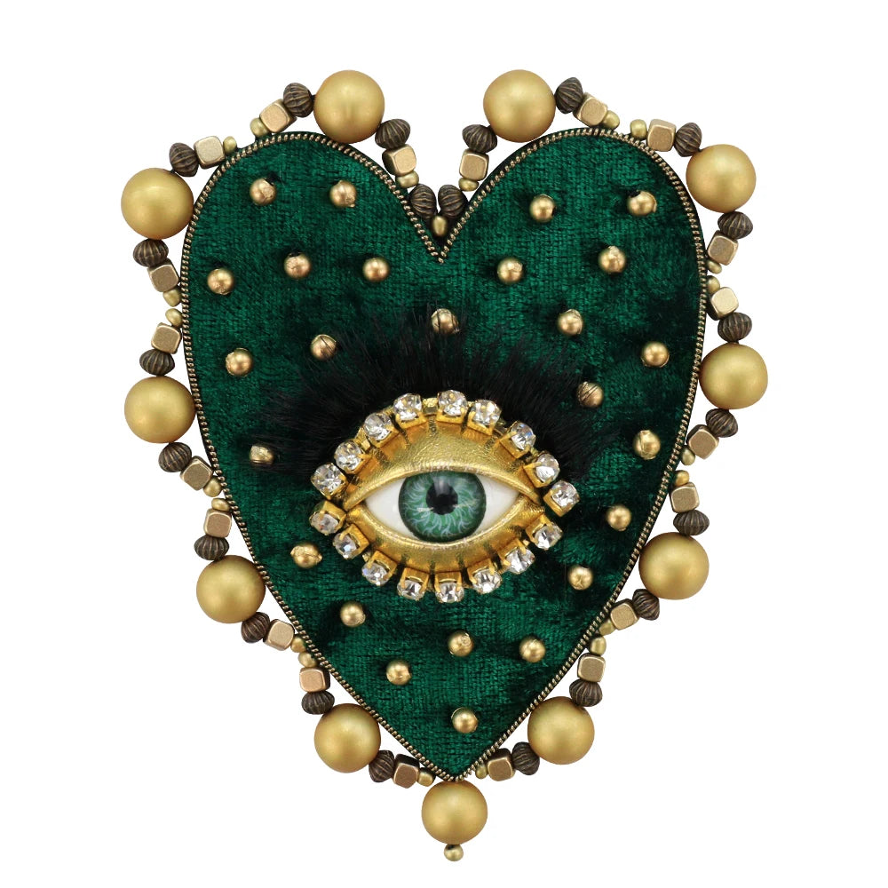 A heart-shaped patch featuring a dark green velvet background, black feathers, and a central gold-colored eye design surround
