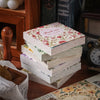 A stack of vintage floral scrapbook paper sheets with pink and green patterns resting on a wooden surface.