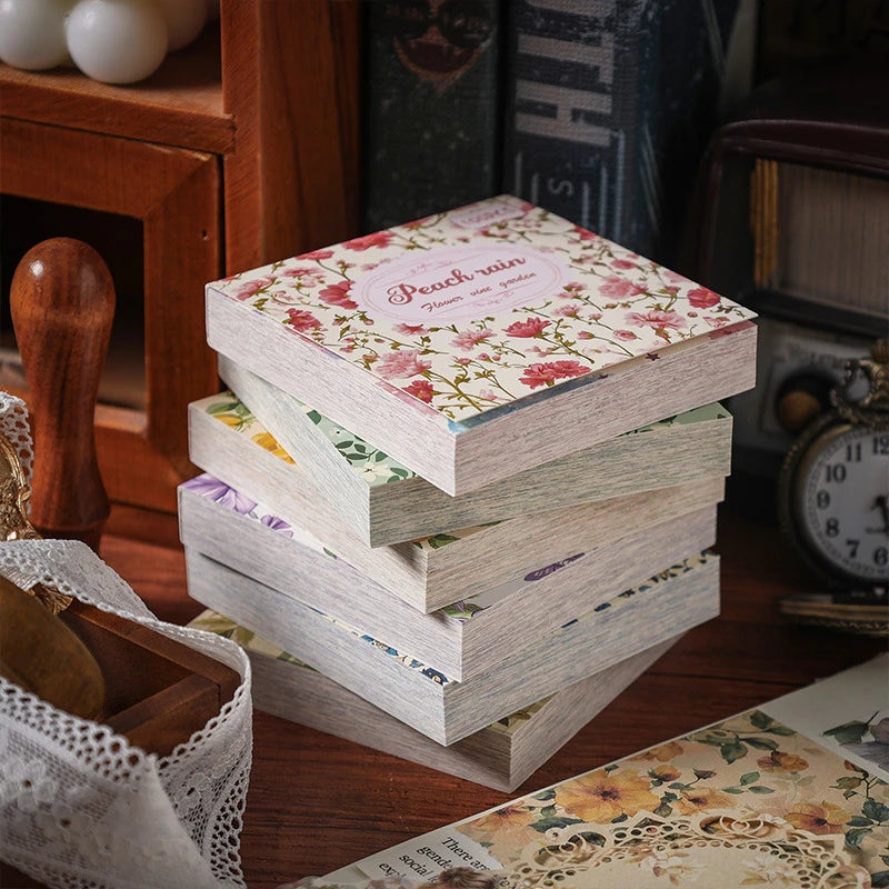 A stack of vintage floral scrapbook paper sheets with pink and green patterns resting on a wooden surface.