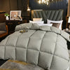 A large gray quilted comforter is displayed on a bed, set against a dark background with a gold chandelier.