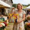 A woman holds a yellow quilted tote bag with colorful birds and floral patterns in an outdoor market setting.