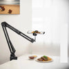 A black adjustable phone stand with an overhead light is positioned above a white table, next to a plate of food.