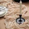 A cream-colored candle in a black holder rests on a beige crinkle gauze table runner next to plates and silverware.