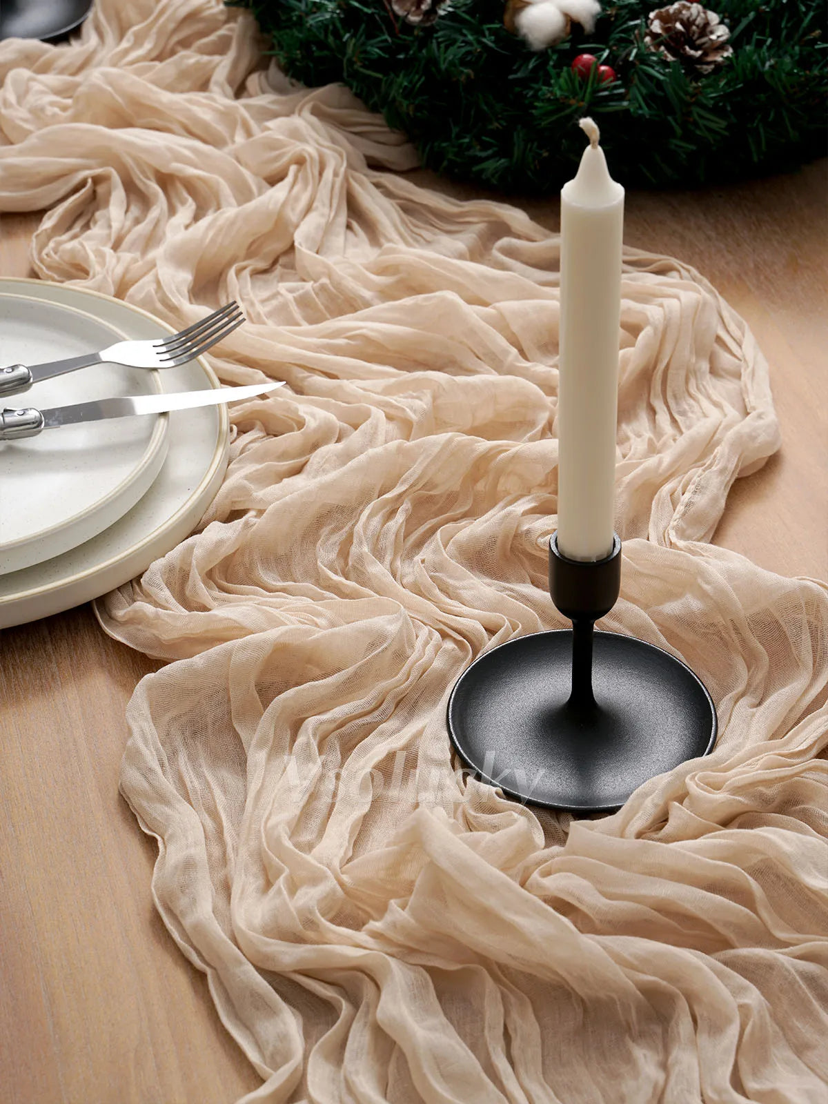 A cream-colored candle in a black holder rests on a beige crinkle gauze table runner next to plates and silverware.
