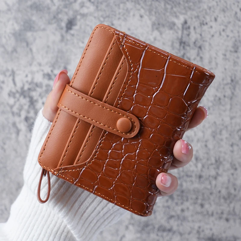 A hand holds a brown crocodile-embossed leather clutch wallet against a gray background.