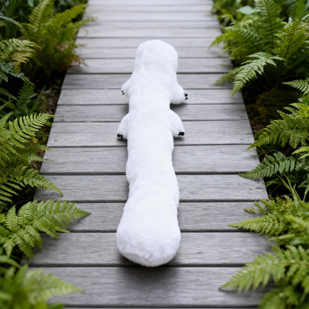 A white plush toy resembling a dog lying on a wooden path surrounded by green ferns.