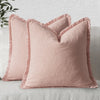 A light pink boho fringe throw pillow cover with a natural linen texture resting on a light gray sofa.
