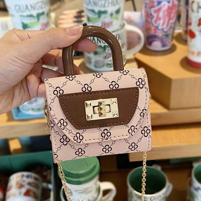 A hand holds a pink mini floral textured crossbody tote bag with brown leather trim and gold chain.