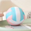 A round simulation volleyball plush toy pillow with pink and light blue stripes resting on a green surface.