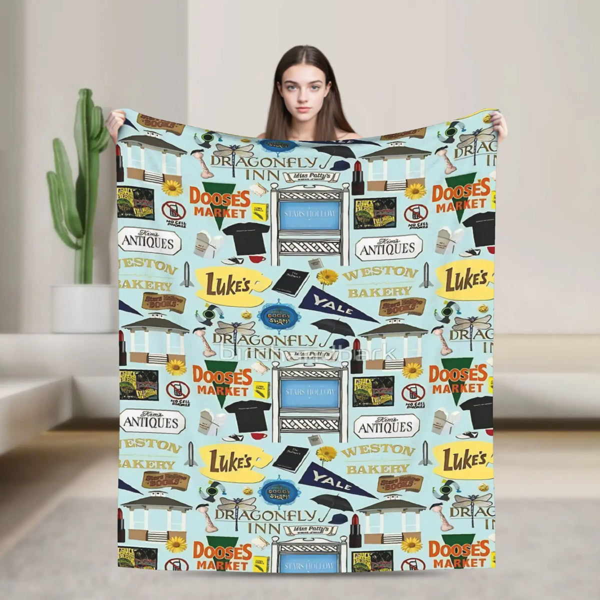 A woman holds up a colorful throw blanket featuring Gilmore Girls themed graphics and text.