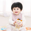 A smiling baby holds a soft plush toy of a cartoon animal with ears and a nose.