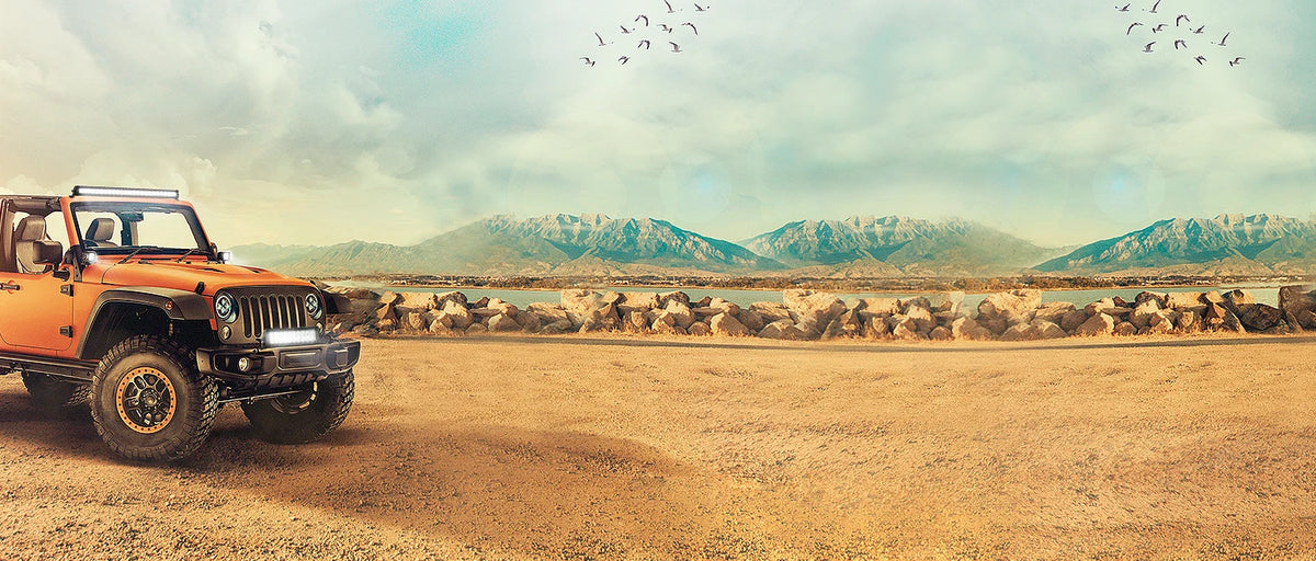 An orange Jeep Wrangler is parked on a dry, dusty terrain with mountains in the background under a cloudy sky.