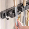 A hand is holding a silver wall-mounted metal mop and broom holder with detachable hooks against a white wall.