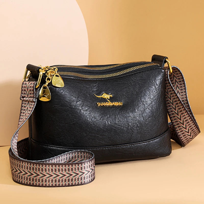 A black PU leather crossbody shoulder bag with gold hardware and patterned strap resting on a beige surface.