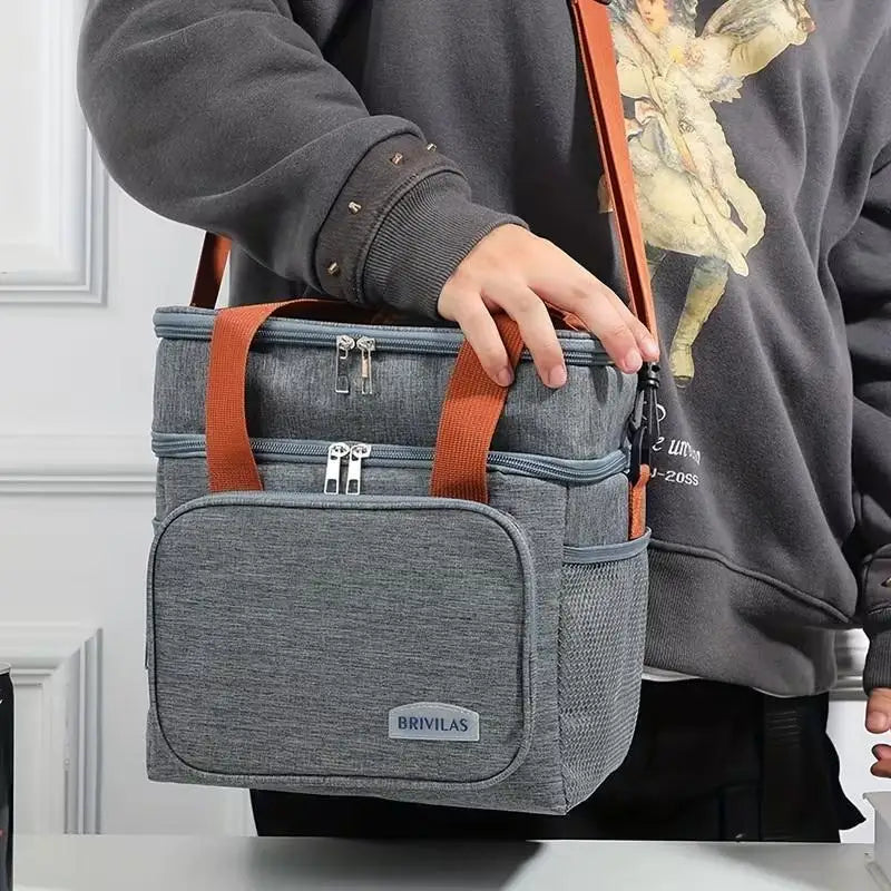 A person holds a gray insulated lunch bag with brown straps and double-layer compartments.