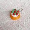 A small, round cake-shaped fidget keychain featuring white frosting and red strawberries with green leaves.