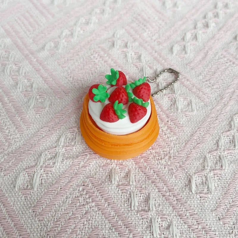 A small, round cake-shaped fidget keychain featuring white frosting and red strawberries with green leaves.