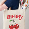 A beige canvas tote bag featuring a red cherry graphic and the text Happy Have a Nice Day.