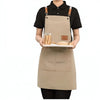 A person is wearing a beige cotton canvas apron with multiple pockets and leather straps over a black shirt.