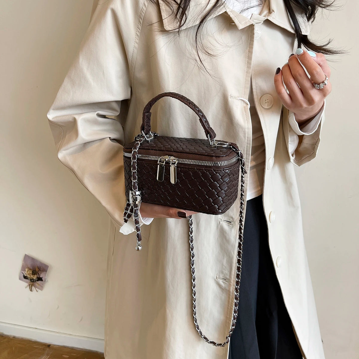 A woman wearing a beige trench coat holds a dark brown woven PU leather crossbody bag with a silver chain strap.