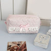 Pink cosmetic pouch with a checkered pattern and white lace trim resting on a light blue surface.