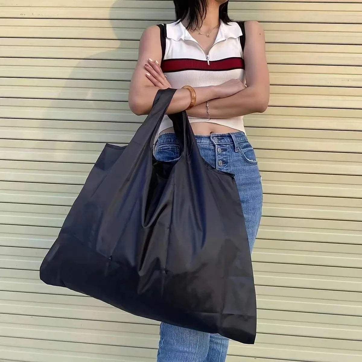 A woman wearing a white top and jeans holds a large black foldable nylon shopping tote bag.