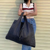 A woman wearing a white top and jeans holds a large black foldable nylon shopping tote bag.