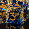 A clear resin polyhedral die with a blue tint and gold markings rests on a reflective surface.