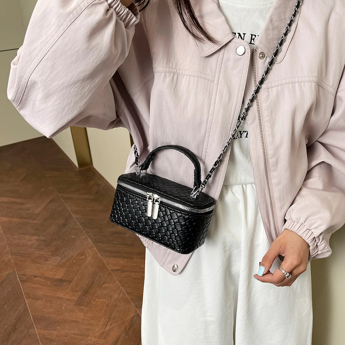 A person wearing a light pink jacket and white top holds a black woven PU leather crossbody bag.