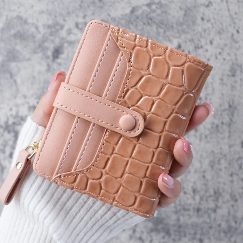 A hand holds a light pink crocodile-embossed clutch wallet with a tan leather texture.