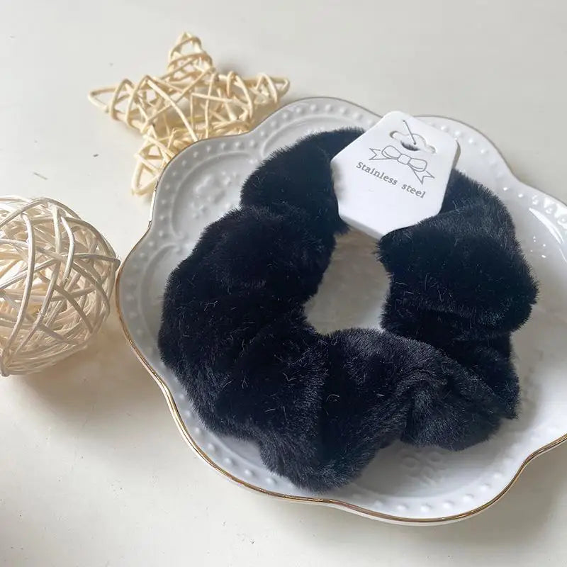 A black plush coil hair tie is displayed on a white plate next to a light beige spherical decoration.