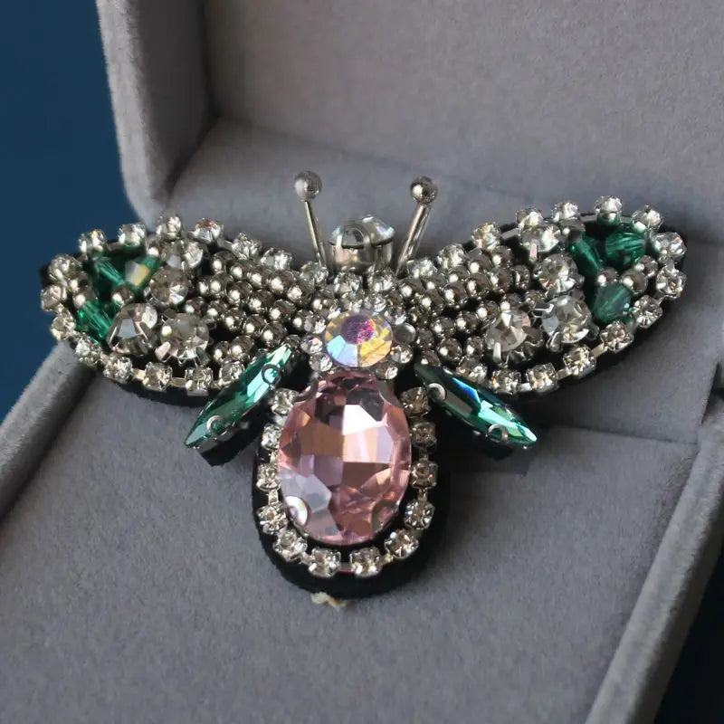 A handcrafted rhinestone bee sew-on patch with sparkling wings and a pink center set against a gray background.
