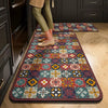 A rectangular, patterned floor mat with a dark border is placed on a light wood floor next to dark cabinetry.