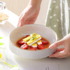 A hand holds a white ceramic soup bowl filled with sliced strawberries and a yellow seasoned rice or salad.