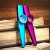 Two silver aluminum kazoo products, one blue and one purple, are displayed on a stack of books.