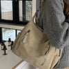 A woman holds a beige vintage matte leather crossbody hobo bag with a gray knit sweater in front of a window.
