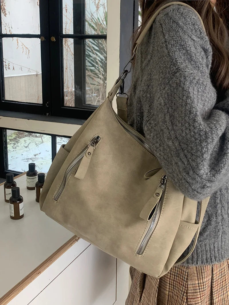 A woman holds a beige vintage matte leather crossbody hobo bag with a gray knit sweater in front of a window.